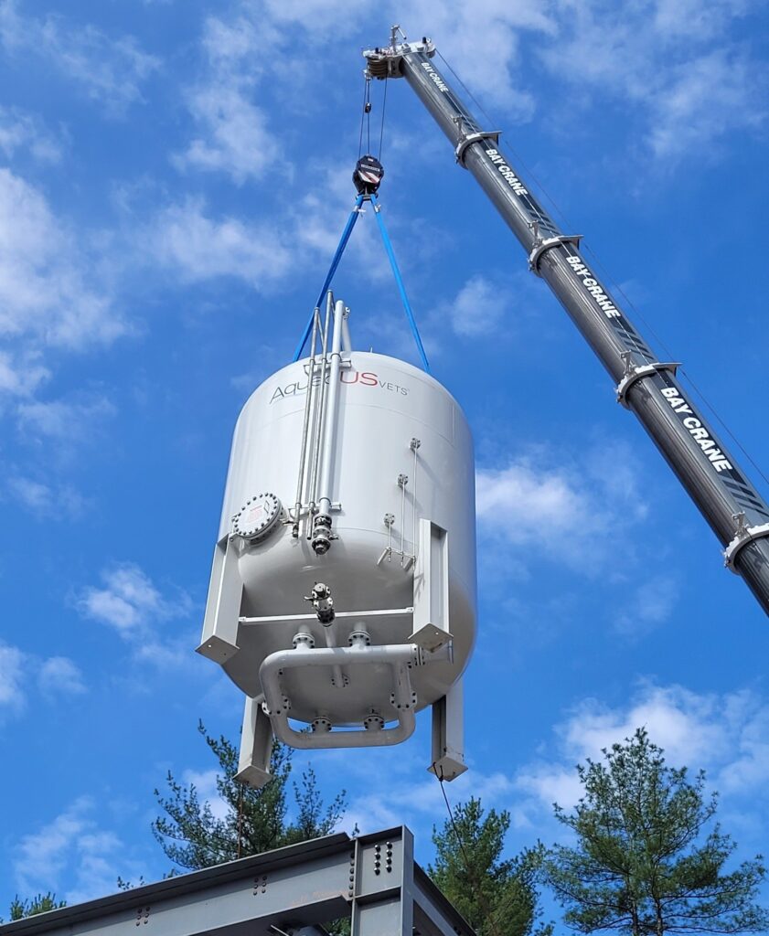 A crane lowers a large tank to the ground with a lovely cloud-dotted sky in the background.