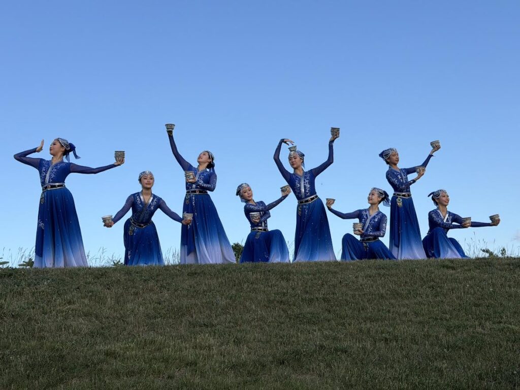 A group of young women, wearing long blue dresses and fancy beaded headdresses dance on the grass. They each hold three rice bowls in various poses. During the dance, they all dance while balancing the rice bowls on their heads. It's impressive!