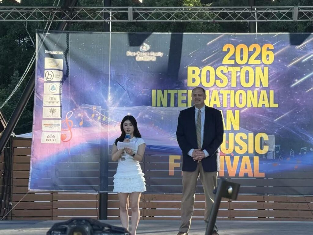 A young woman in a white dress reads from a paper while a tall man in a suit stands next to her. On the background, the stage back reads "2026 Boston International Asioan Music Festival."
