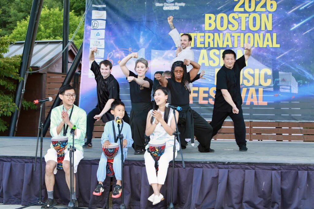 A couple and a young child sit on the edge of the stage, all holding colorful drums between their legs. In the background, a group of people, mostly wearing black, strike a martial arts pose.