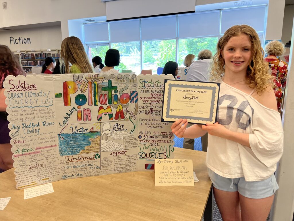 A blonde girl stands in front of a poster that says, in multi-colored letters Pillution in MA. The poster has a lot of other information on it.