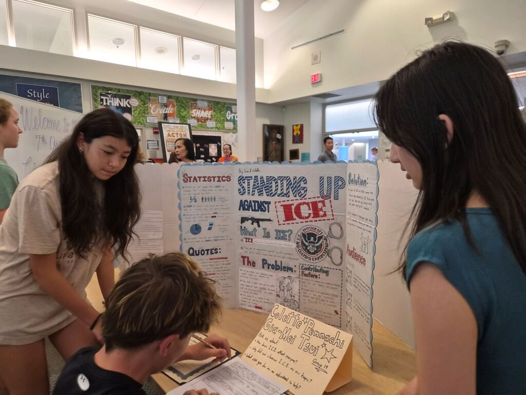 Two girls stand by a poster where the largest text says Standing Up Against ICE. A third kid is sitting in front of the poster filling out a form.