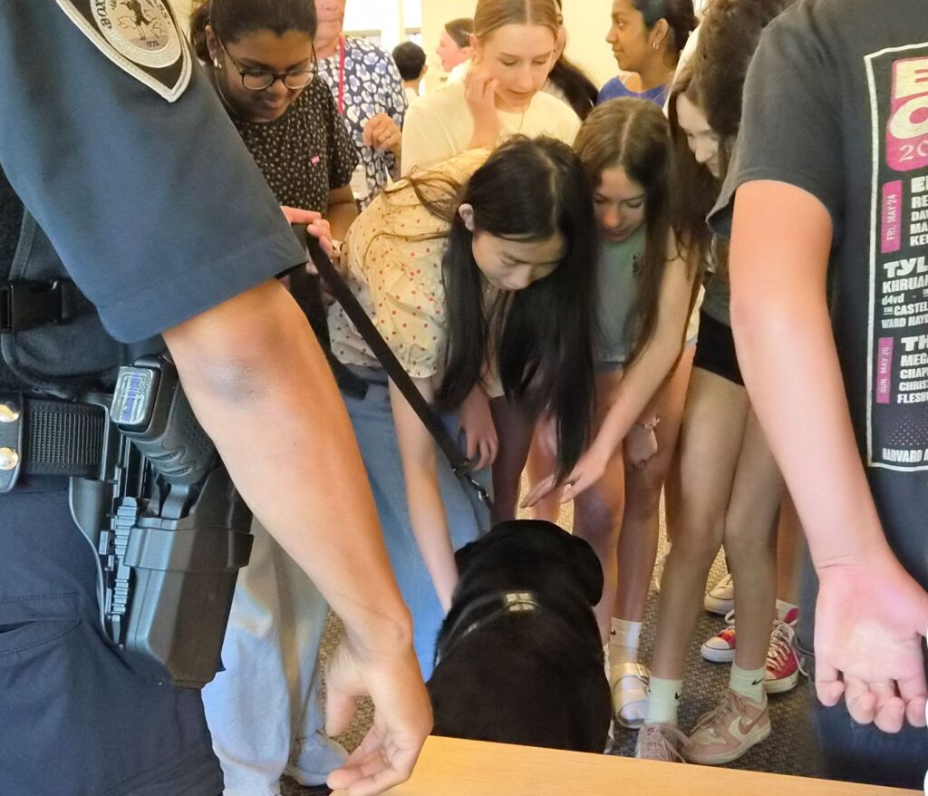 A black lab dog is surrounded by kids reaching out to pet him. In the foreground, a police officer stands, gun in holster.