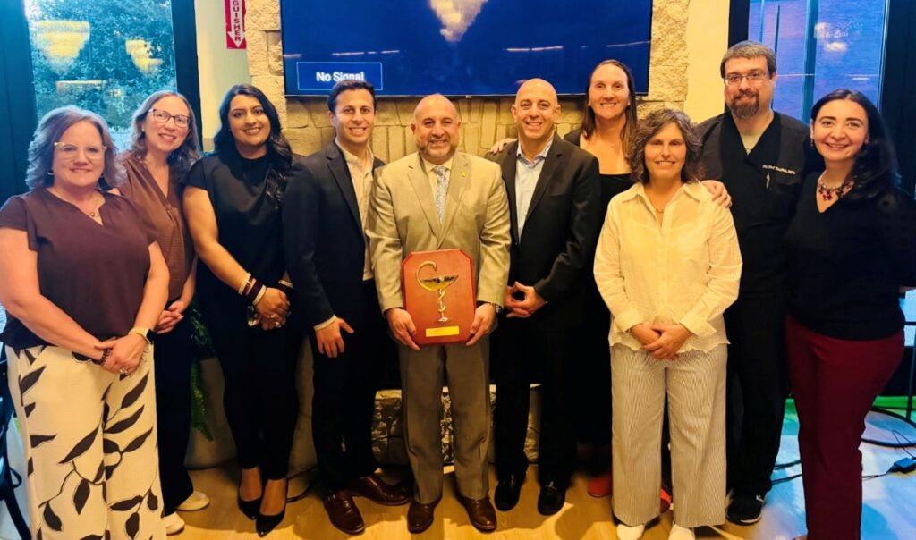 In the middle of the photo a balding man in a suit holds a plaque that has a stemmed bowl. Surrounding him are a several other people (employees, this time).