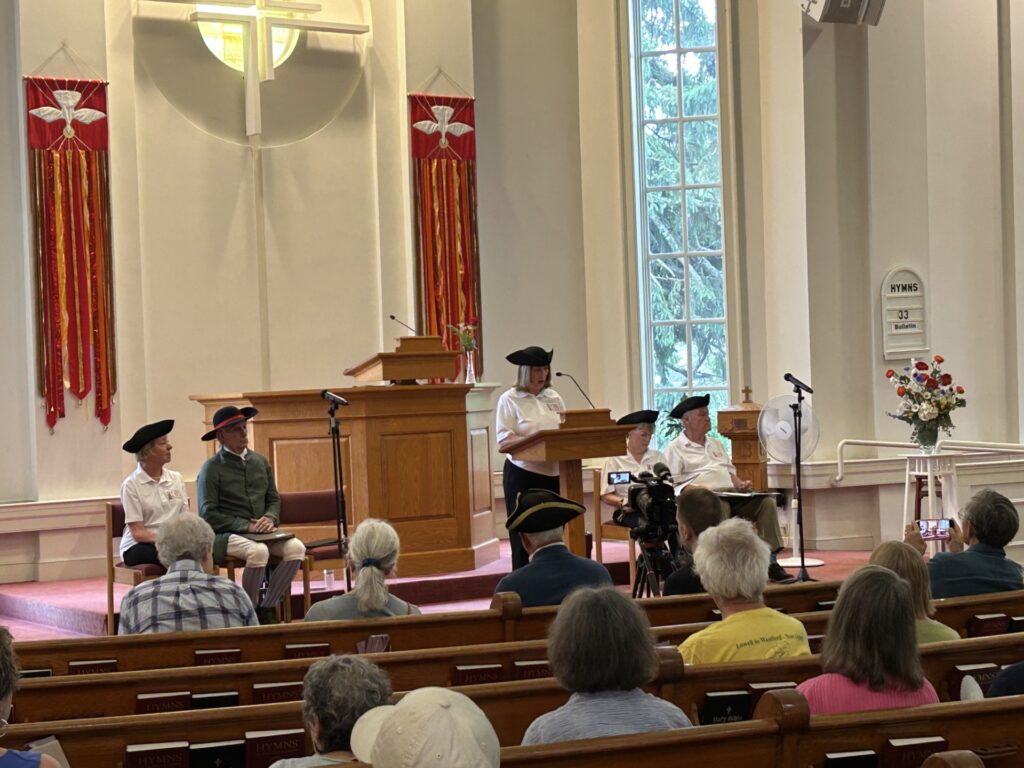 A woman wearing a tricorner hat (and an Acton 250 polo) reads at the podium. The audience listens from the pews.