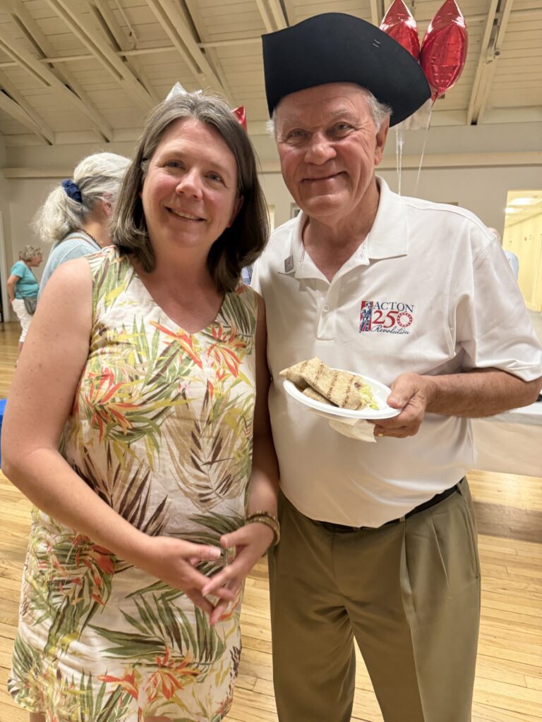 A woman in a flowered dress stands next to an older man wearing a tricorner hat and Acton 250 polo shirt. He's holding a plate of food.