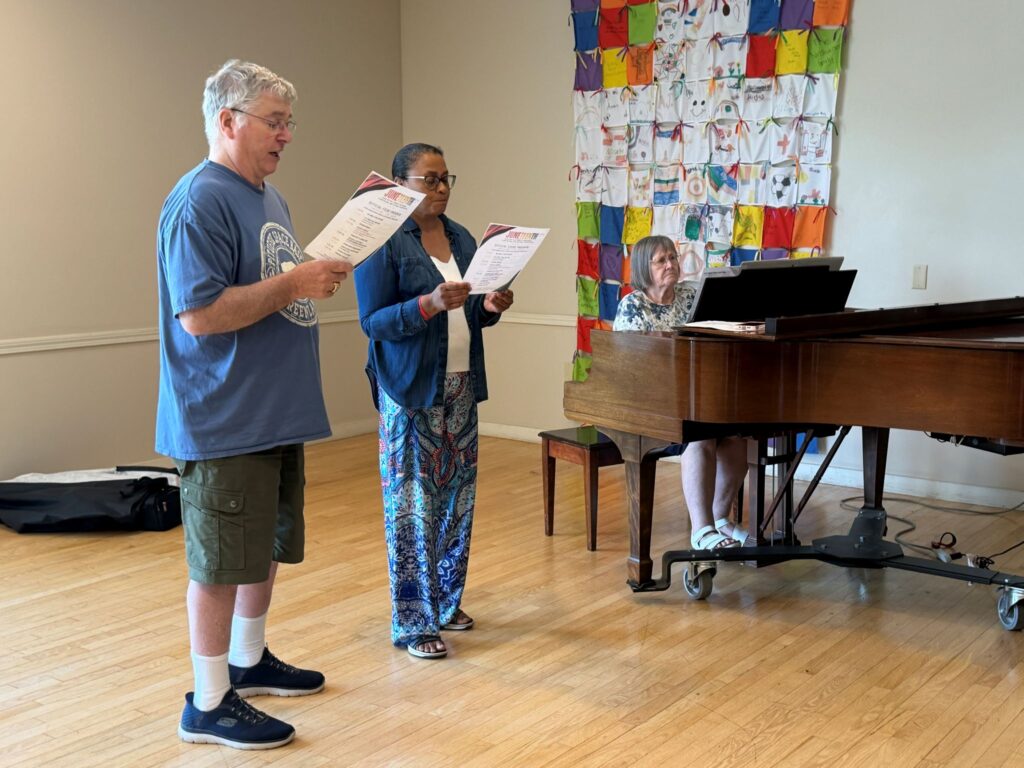 A grey-haired white man and a black woman stand next to an older woman playing a piano. The singers are holding a paper that may contain the lyrics to "Lift Every Voice and Sing."