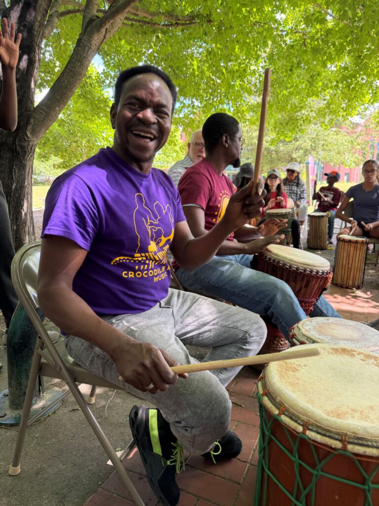 In the foreground, a smiling Black man plays a drum. He's wearing a bright purple Crocodile River t-shirt. In the background are two drummers on the left, and a bunch of folks playing drums under a tree.