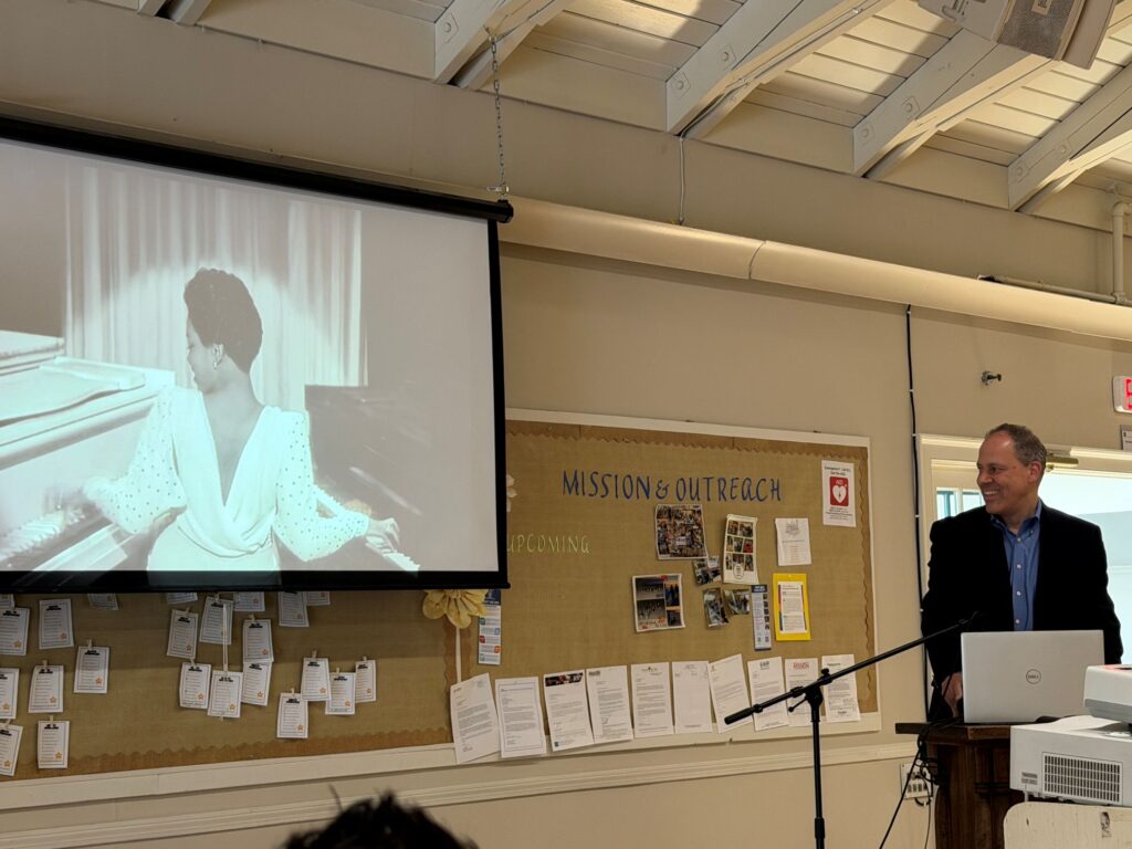 A man stands at a podium, Behind him, a screen shows an elegantly dressed Black woman sitting in between two pianos.