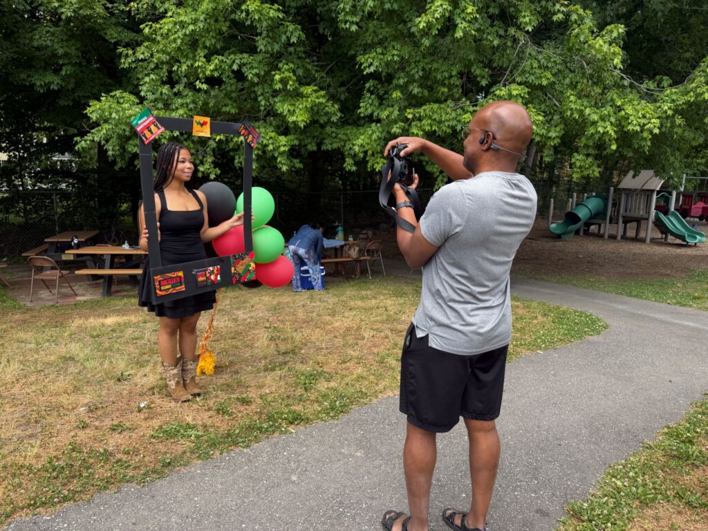 A young woman has her picture taken. She is holding a large black frame that has colorful objects glued on.