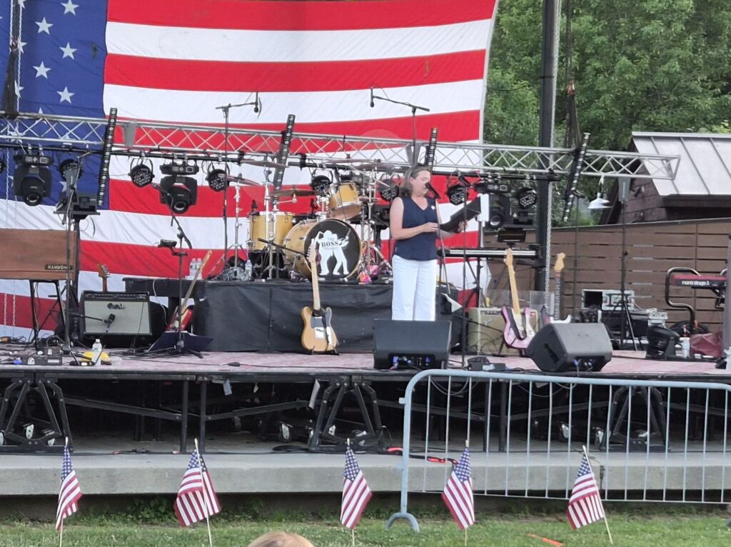 Acton Select Board Chair Alissa Nicols standing on stage in front of a large American Flag in Nara Park in Acton.