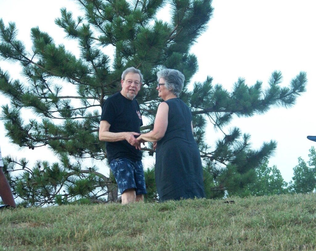 A man and a woman holding hands and smiling on a hill in front of a large pine tree in Nara Park in Acton.