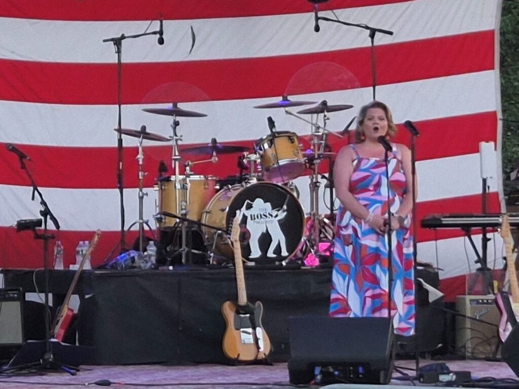 Singer Kiki Donohue on a stage at Nara Park in front of a large American Flag.