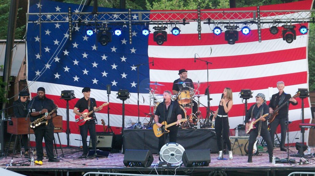 The Boss Project on stage at Nara Park in Acton with instruments and microphones, and a large American Flag in the background.