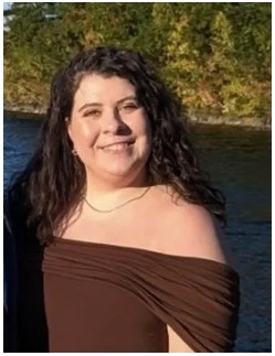 Ellie Anderson in a brown off the shoulder dress smiling with a hill full of trees in the background.