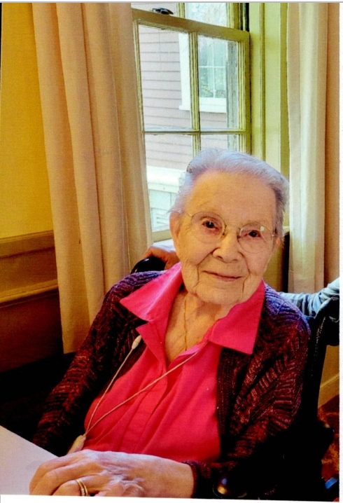 Lillian Jaehnig with glasses and her hair in bun, wearing a red shirt with a dark sweater seated in front of a window with long curtains.