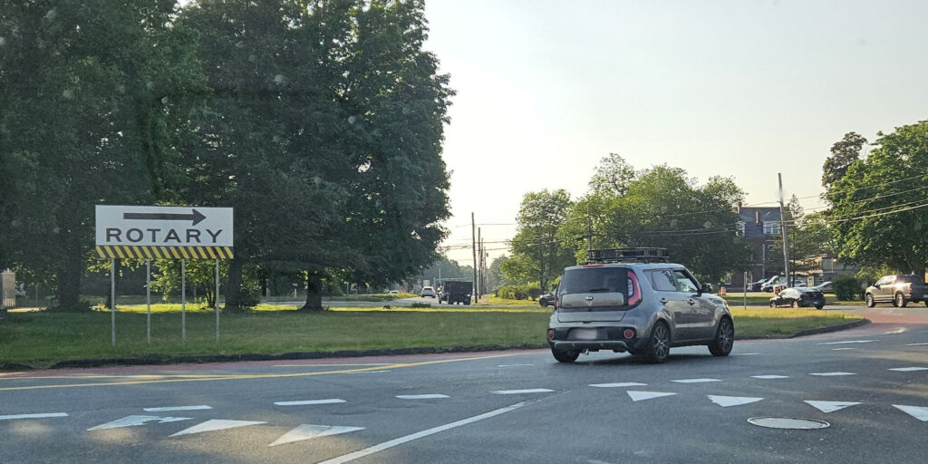 Three cars driving through the rotary on Route 2 in Concord, MA, around the center island with green grass and large green trees.